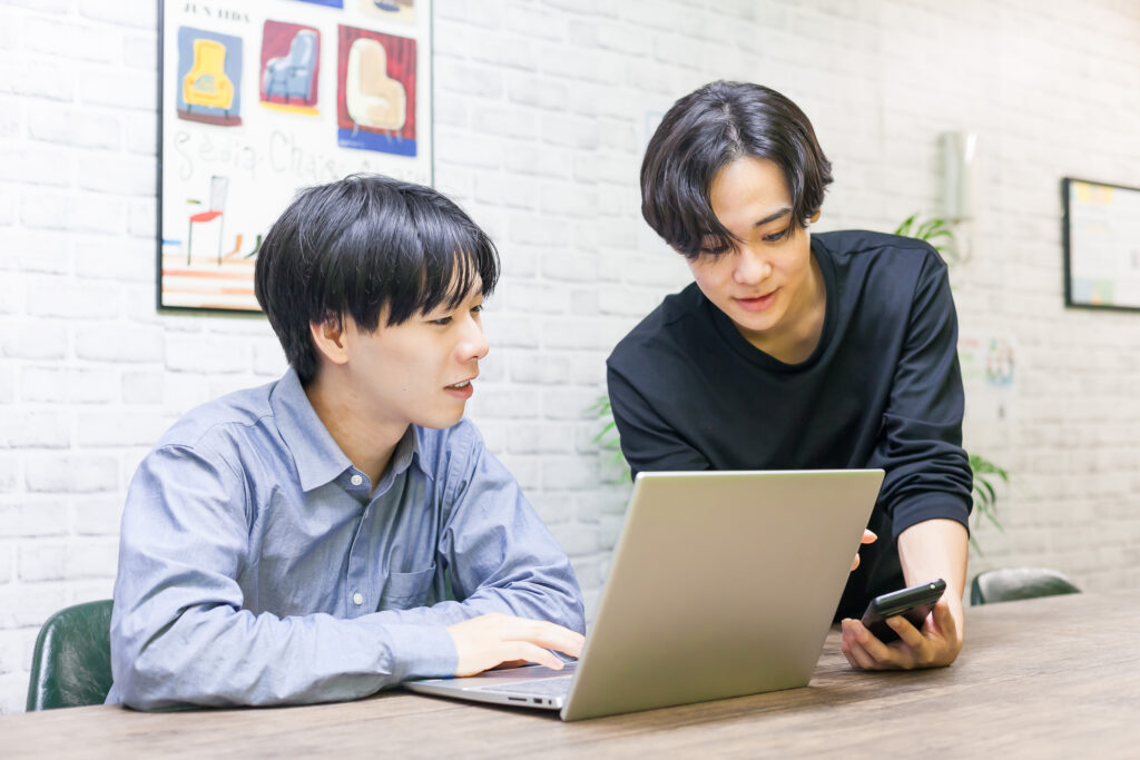 男性2人が事務室でパソコンとスマホを見ながら一緒に作業をしている画像。