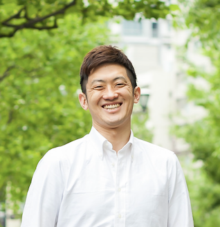 代表理事の高橋が、外で緑の木を背景に笑顔で立っている画像。