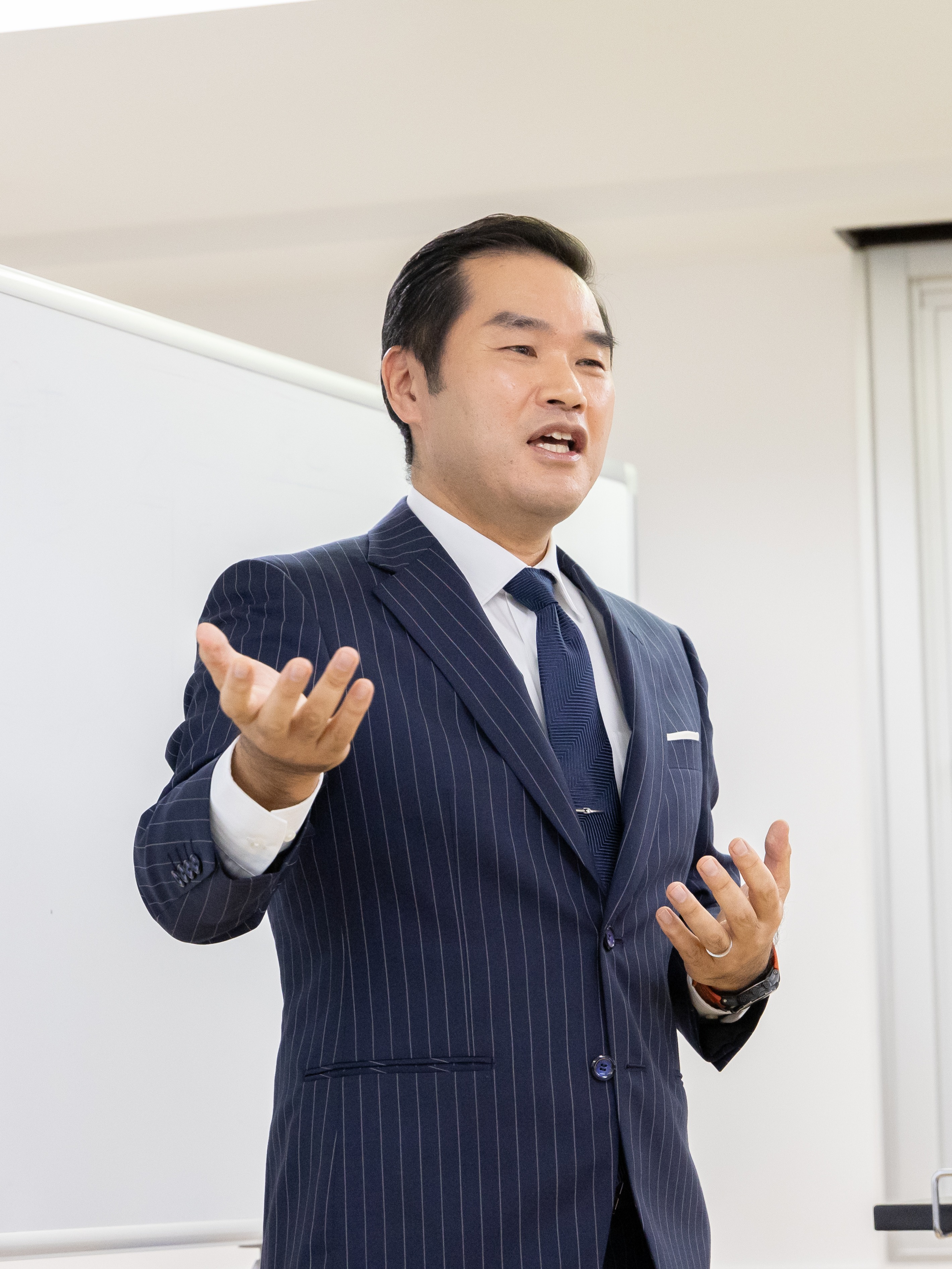 寺田がスーツ姿でホワイトボードの前でプレゼンをしている画像。